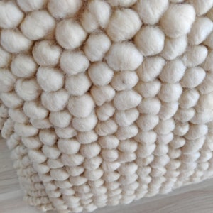 May include: Close-up of a cream-colored throw pillow with a textured, puffy surface. The pillow is made of a soft, woven material with a unique, bumpy design.