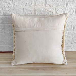May include: A cream-colored throw pillow with a white pom-pom trim. The pillow is made of a soft, woven fabric and has a zipper closure.
