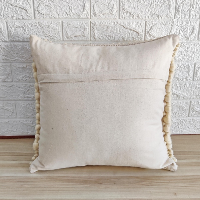 May include: A cream-colored throw pillow with a white pom-pom trim. The pillow is made of a soft, woven fabric and has a zipper closure.