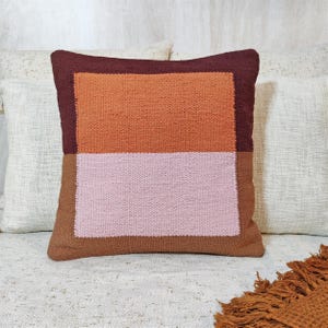 May include: Square cushion with a geometric design. The cushion features a rectangular pattern in shades of brown, orange, and pink. The cushion is made of woven fabric and is placed on a white surface.