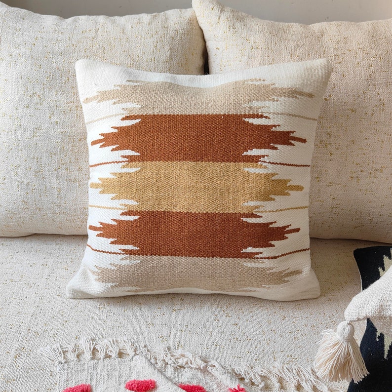 May include: A white throw pillow with a geometric pattern in shades of brown and beige. The pillow has a woven texture and a simple, modern design.