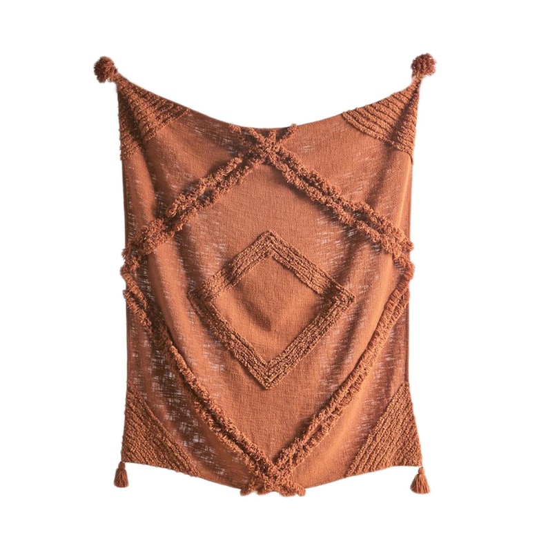 May include: A terracotta-colored throw blanket with a diamond pattern and fringe trim. The blanket has a textured woven design and tassels on the corners.