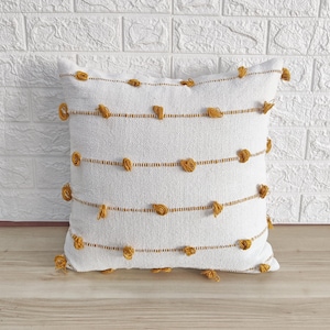 May include: White throw pillow with a woven texture and yellow tassels. The pillow has a subtle striped pattern and is decorated with small yellow pom-poms.