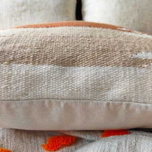 May include: Close-up of a beige and white woven throw pillow with a brown stripe. The pillow has a plain beige fabric backing.