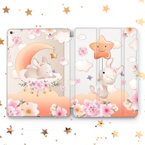 May include: A tablet case with a cute design featuring two bunnies, one sleeping on a crescent moon and the other holding a star balloon. The case has a pink and white floral pattern with gold stars.