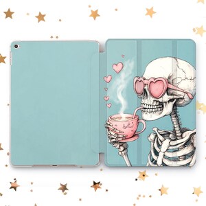 Könnte beinhalten: Eine blaue und weiße iPad-Hülle mit einem Skelett, das eine rosafarbene Herz-Sonnenbrille trägt und aus einer rosafarbenen Teetasse trinkt. Das Skelett ist von rosa Herzen umgeben.