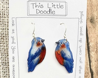 May include: A pair of bluebird earrings with red and white accents. The earrings are made of resin and have silver hooks. The card reads "This Little Doodle" and "Who says you can't do great things!" with the artist's name "Sarah Katzel" at the bottom.