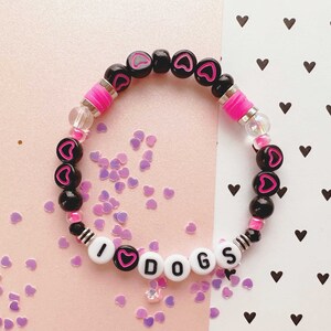 May include: A black and pink beaded bracelet with the words "I love dogs" spelled out in white beads. The bracelet is on a pink and white background with small hearts.