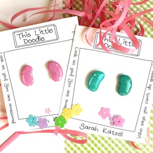 May include: Two cards with the text "This Little Doodle" and "Look at you! You are awesome!" and "Who says you can't do great!"  The cards have jelly bean shaped buttons in pink and teal with glitter. The cards are decorated with small paper flowers in various colors.