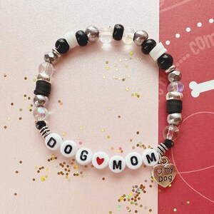 May include: A black and white beaded bracelet with the words "DOG MOM" spelled out in white beads with a red heart bead between the words. A silver charm with the words "my dog" is attached to the bracelet.