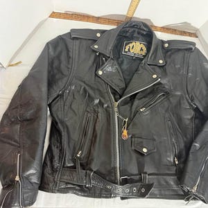 May include: A black leather motorcycle jacket featuring a notched lapel, silver-tone metal zippers, and a waist belt with a buckle. The interior label reads FORCE GENUINE LEATHER. The jacket includes shoulder epaulets, zippered cuffs, and pockets.