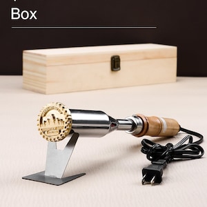 May include: A gold-colored branding iron with a wooden handle and a metal stand. The iron has a circular design with a crown and the words "Certified Organic" and "Original" around the edge. The iron is plugged into an electrical outlet and is ready to use. The iron is also included in a wooden box.