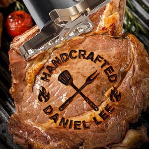 May include: A close-up of a grilled ribeye steak with a branding iron. The branding iron is silver and black, and the steak has the words "HANDCRAFTED DANIEL LEE" branded on it. Other grilled items are visible.