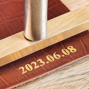 May include: A metal tool stamps the date "2023.06.08" onto a textured, reddish-brown leather surface. The tool includes a rectangular brass bar and a cylindrical metal piece. The leather has a crocodile-like pattern. The image is a close-up.