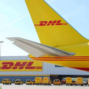 May include: A yellow DHL airplane with the DHL logo on the tail. The airplane is parked in front of a building with the DHL logo on the side. There are several yellow DHL delivery vans parked in front of the building.