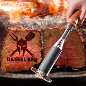 May include: A close-up of a branding iron with a wooden handle being used to brand a piece of meat with the words "DANIEL BBQ" and a graphic of a knight's helmet with crossed swords and a fork and spatula.