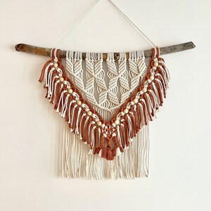 Peut inclure: Une suspension murale en macramé avec une branche de bois en haut. Le macramé est fait de fil blanc et marron et a des perles de bois tissées dans le motif. La suspension a une frange en bas.