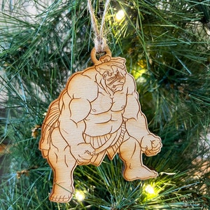 Puede incluir: Un adorno navideño de madera con forma de luchador de sumo. El adorno está pintado en tonos marrones y presenta un diseño detallado de los músculos y la ropa del luchador.
