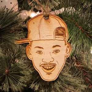 May include: A wooden Christmas ornament featuring a cartoon portrait of Will Smith wearing a baseball cap. The ornament is brown and has a string for hanging.