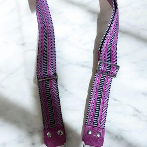 May include: A patterned bag strap with a pink, black, and white design. The strap has silver-colored metal hardware, including clasps and buckles. The ends are finished with textured, dark pink leather.