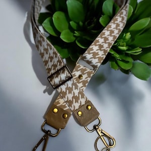 May include: A tan and white houndstooth patterned bag strap with gold-colored metal hardware. The strap features a leather-like material at the ends with rivets and swivel clasps. A small key charm with the word "DREAM" hangs from one clasp.