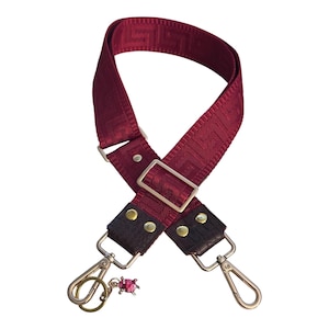 May include: A deep red adjustable bag strap featuring a woven Greek key pattern. The strap includes dark brown faux leather ends secured with gold-tone metal rivets, two gold-tone lobster claw clasps, and a small pink ladybug charm attached.