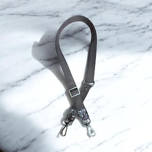 May include: A gray crossbody bag strap with silver-tone metal hardware. The strap has a patterned accent near the clips. The strap is made of a durable fabric and features a metal buckle for adjustment. The background is a white marble surface.