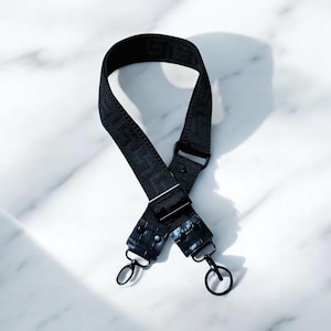 May include: A black adjustable bag strap featuring a woven geometric pattern throughout the fabric. The strap includes black metal hardware, including sliding buckles and two circular swivel clasps attached to textured black faux leather ends.