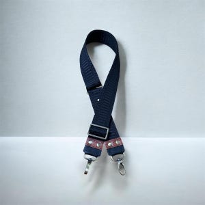 May include: A navy blue patterned bag strap with silver-tone metal hardware. The strap has a textured design and is accented with pink leather details and silver rivets. The strap features two silver swivel clasps.