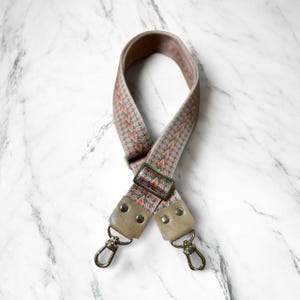 May include: A patterned bag strap with a geometric design in shades of gray, orange, and green. The strap has tan leather accents and metal hardware, including a buckle and swivel clasps. The strap is approximately 40 inches long.