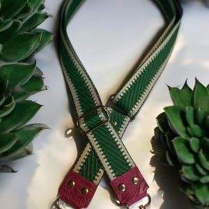 May include: Green and cream-colored patterned bag strap with gold-tone hardware. The strap features a woven design with green and cream stripes. The ends are a deep pink leather with gold rivets and clasps. The strap is designed to be attached to a bag.