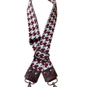 May include: A burgundy and white houndstooth patterned bag strap with a leather accent. The strap has metal hardware, including a buckle and swivel clasps. The strap is designed to be adjustable and attachable to a bag.