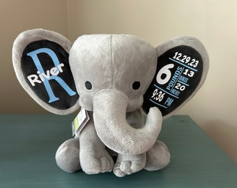 May include: Gray plush elephant with blue and white details. One ear has the name "River" and the other ear has the date "12.29.23", time "9:36 PM", weight "6 pounds", and length "20 inches".