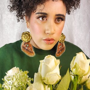 May include: A person wearing a green shirt with butterfly and flower earrings. The person has a nose ring and is surrounded by white roses.