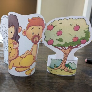 May include: Paper cutouts of biblical figures and an apple tree. The figures include a man with a lion, a man with a beard, and a figure in black and white. The apple tree has red apples and green leaves. The cutouts are on a wooden surface.