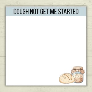 以下が含まれることがあります： 黒い文字で「DOUGH NOT GET ME STARTED」と書かれた白いメモ帳。メモ帳には、パンとサワードウスターターの瓶の漫画イラストが描かれています。メモ帳は明るい木目調の背景にあります。