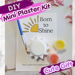 May include: A DIY mini plaster kit featuring a white sun-shaped plaster figure, a blue-handled paintbrush, and two small paint pots containing yellow and orange paint. A white card displays the text Born to Shine and more creative fun at paperscissorscraft.