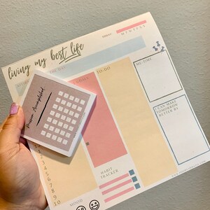 May include: A pink and yellow daily planner with the text "living my best life" at the top. The planner has sections for affirmations, goals, to-do list, me-time, habit tracker, and mood. There is a small calendar sticker on the planner.