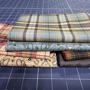 May include: A stack of fabric pieces with various patterns. The fabrics include plaid, paisley, and western-themed designs in shades of blue, tan, red, and gray. The fabrics are neatly folded and stacked on a blue cutting mat.