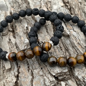 May include: Two bracelets, one with black lava rock beads and the other with tiger's eye beads. The bracelets are stacked on top of each other on a wooden surface.