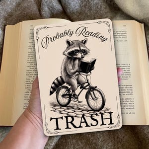 May include: A black and white illustration of a raccoon wearing glasses and riding a bicycle while reading a book. The sign reads "Probably Reading Trash".