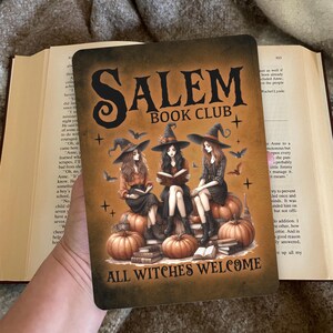 Op de afbeelding: Een rechthoekig bord met de tekst "Salem Book Club" en "All Witches Welcome". De afbeelding toont drie vrouwen met heksenhoeden die op pompoenen zitten en boeken lezen, met vleermuizen die om hen heen vliegen. De achtergrond is een verloop van bruin en oranje.