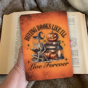 Op de afbeelding: Oranje rechthoekige boekenlegger met Halloween-thema. Het toont een skelet met een heksenhoed dat leest, met pompoenen, boeken en de tekst "Buying Books Like I'll Live Forever".
