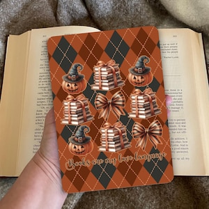 Op de afbeelding: Een rechthoekig decoratief item met een herhalend patroon van oranje en zwarte ruiten. Het ontwerp bevat afbeeldingen van pompoenen met heksenkostuums, stapels boeken en strikken. De tekst "Books are my love language" is ook aanwezig.
