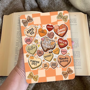Op de afbeelding: Een rechthoekige stickervel met een oranje en witte geruite achtergrond. De vel bevat hartvormige stickers met herfstthema-zinnen zoals "Pumpkin Spice" en "Sweater Weather". Decoratieve strikaccenten zijn ook aanwezig.