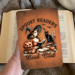 Op de afbeelding: Een rechthoekig oranje bord met de tekst "Spooky Readers Book Club" toont een heks die een boek leest, een zwarte kat, pompoenen en vleermuizen. De illustratie heeft een Halloween-thema.