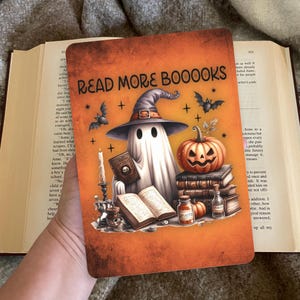 Op de afbeelding: Oranje Halloween-thema boekomslag met de tekst "READ MORE BOOOOKS". De illustratie toont een spook met een heksenhoed, een uitgesneden pompoen, vleermuizen, boeken en kaarsen. Een griezelig en leuk ontwerp.