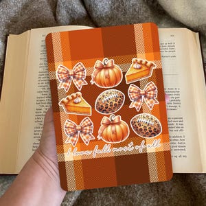 Op de afbeelding: Een herfstthema notitieboek met een oranje en bruin geruit patroon. Het heeft stickers van pompoenen, strikken, American footballs en taartpunten. De tekst "I love fall most of all" is zichtbaar.
