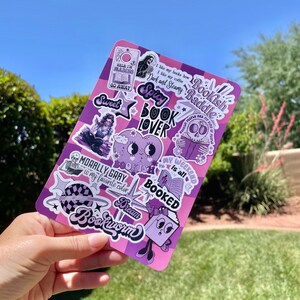 May include: A pink and purple sticker sheet with various bookish designs. The stickers feature phrases like "Bookish Daddie", "Spicy Book Lover", "Morally Gray is my favourite colour", and "Dream Bookworm".