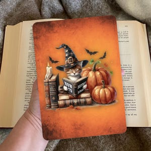 Op de afbeelding: Oranje rechthoekige plaat met een katje met een heksenhoed die een boek leest met de titel "How to Destroy Humans". Pompoenen, vleermuizen en een kaars zijn ook afgebeeld.